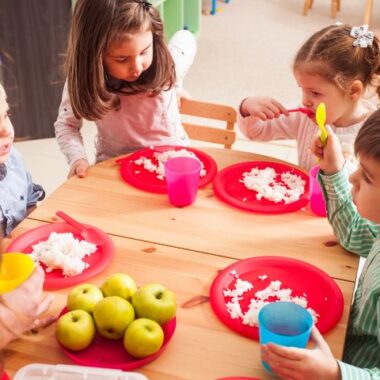 Dzieci jedzą przy stole swoje posiłki, a mama zachęca je do spożywania.
