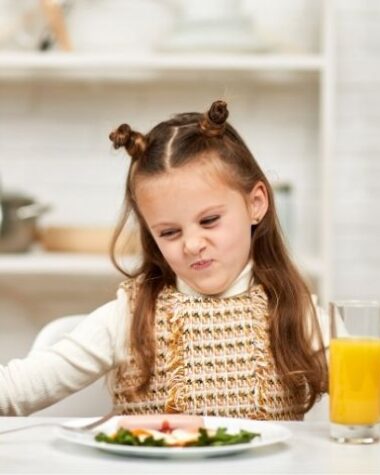 Jak długo dziecko może nie jeść? Na zdjęciu dziewczynka przy stole z grymasem na twarzy.